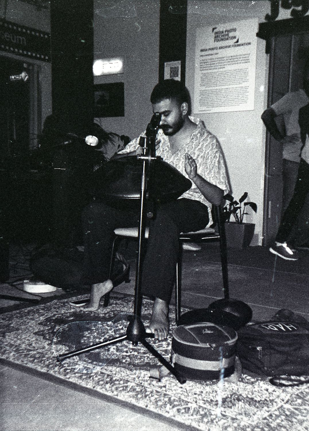 Grainy black-and-white photo of Ashim Bery playing a handpan on a rug, a microphone in front of him and exhibition posters behind