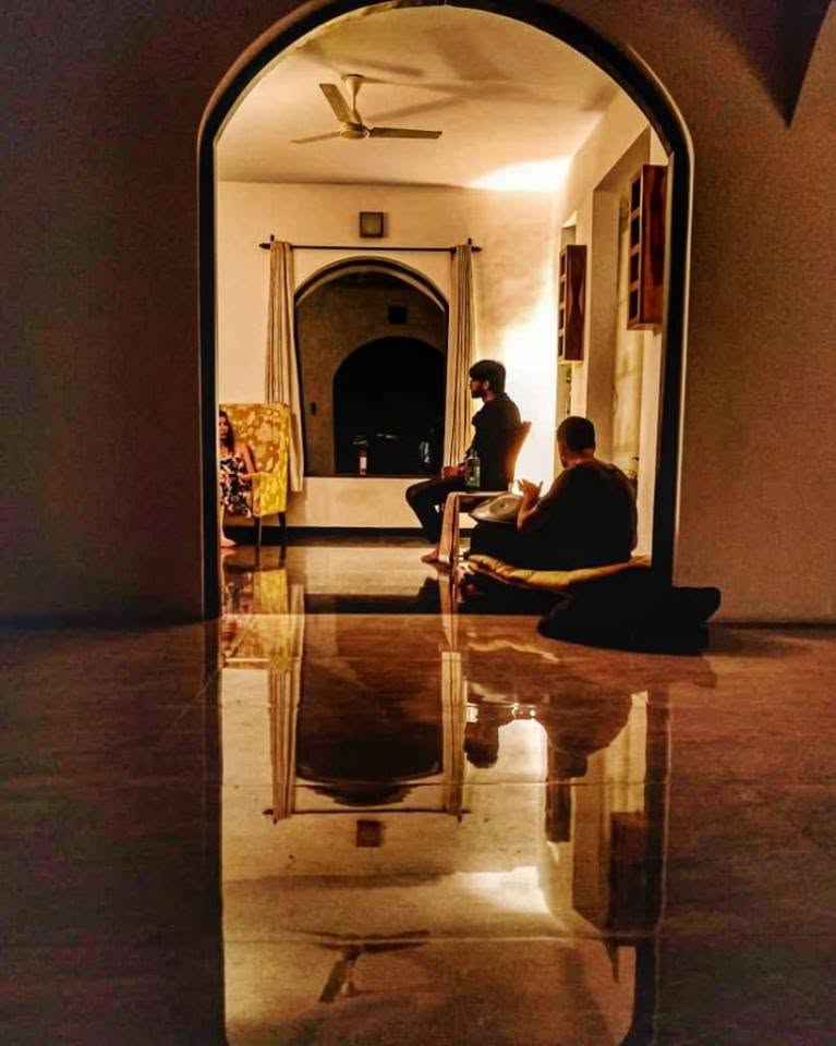 Seen through an archway across a reflective floor at Mandrem House, Goa, Ashim Bery plays a handpan beside an actor in a warmly lit room