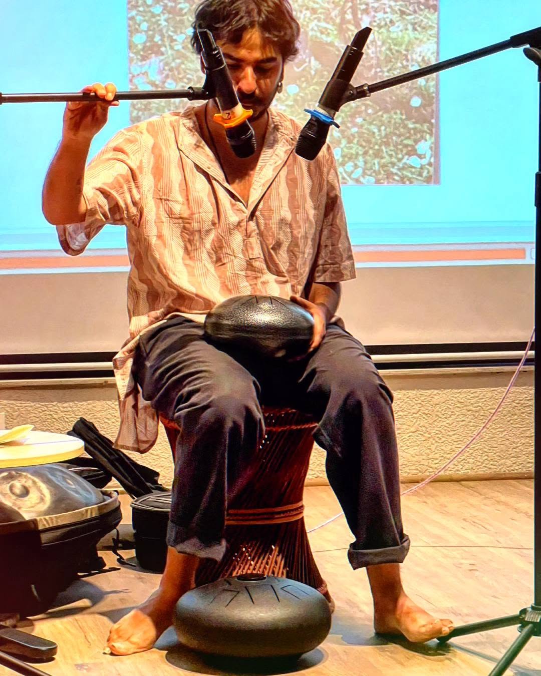Ashim Bery seated barefoot on a cane stool playing a small steel drum, a projection behind and a handpan in its case at his feet