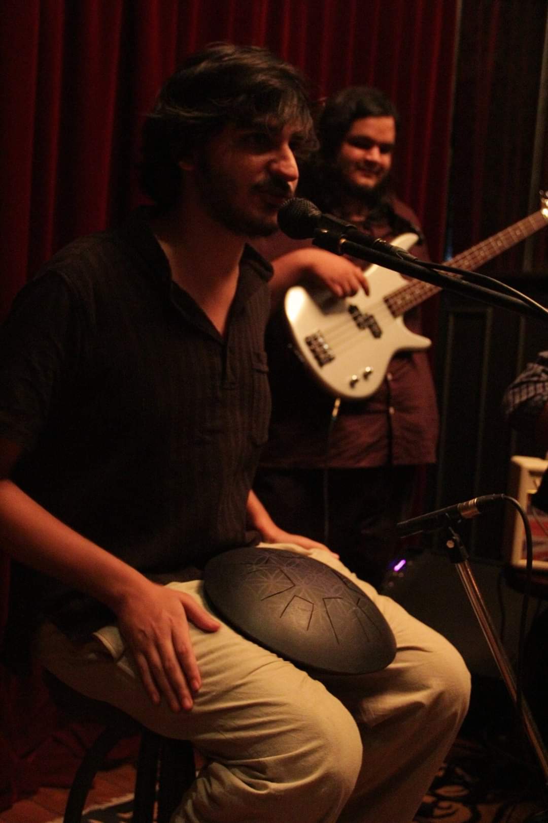 Ashim Bery with a tongue drum on his lap, speaking into a microphone, a bass player smiling behind him