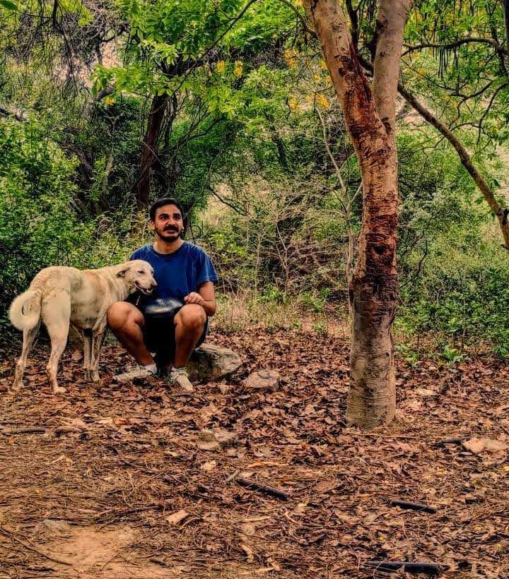 Ashim Bery crouching on a leaf-strewn forest floor with his handpan, a pale dog leaning in beside him, trees all around