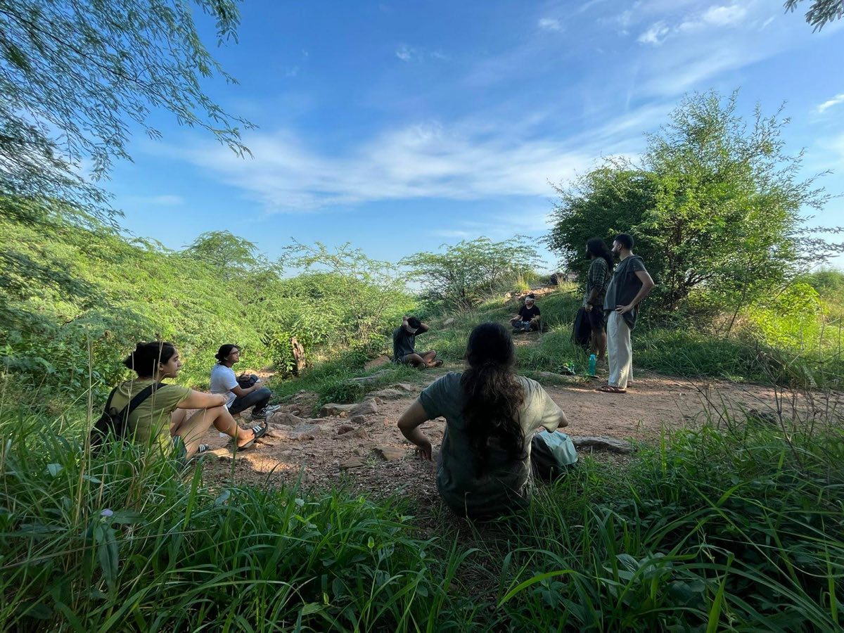 Walkers sitting on a grassy rise under a blue sky, some wearing blindfolds, while others stand nearby