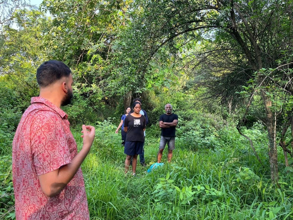 Ashim Bery in a patterned shirt speaking to walkers who stand with their eyes closed in tall grass