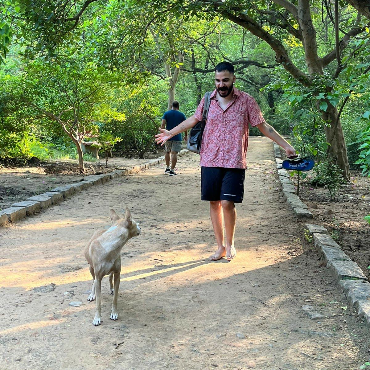 Ashim Bery walking barefoot on a sunlit forest path, laughing as he greets a dog, his sandals in hand