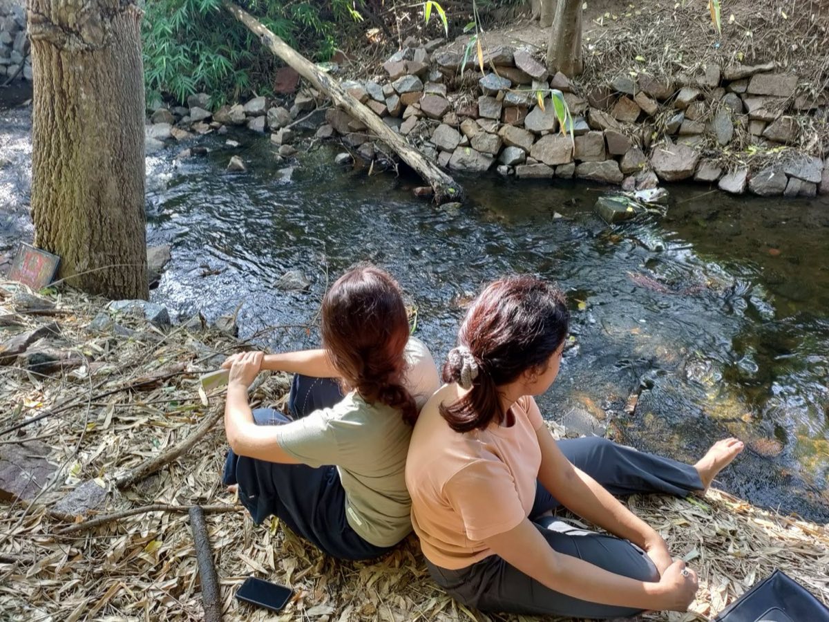 Two walkers sitting back to back on fallen leaves beside a clear stream